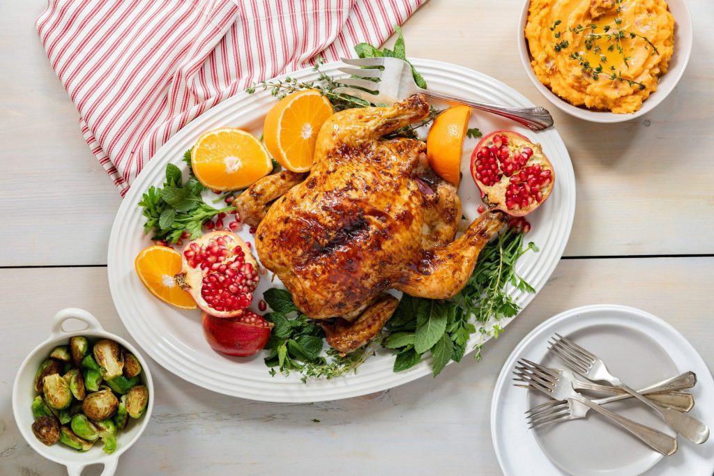 Whole chicken served on white plate with greens, pomegranates, and oranges